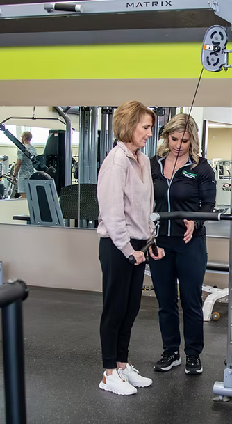 Two women standing together in a gym, looking at a weight machine.