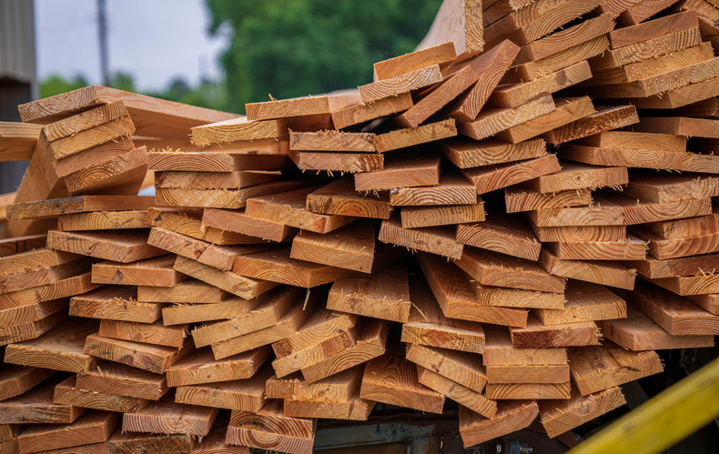 Pile of rough-cut lumber; tan wood planks stacked haphazardly outdoors.