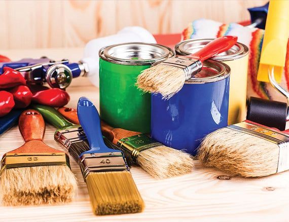 Paint cans and brushes of various colors on a wooden surface, with a paint sprayer in the background.
