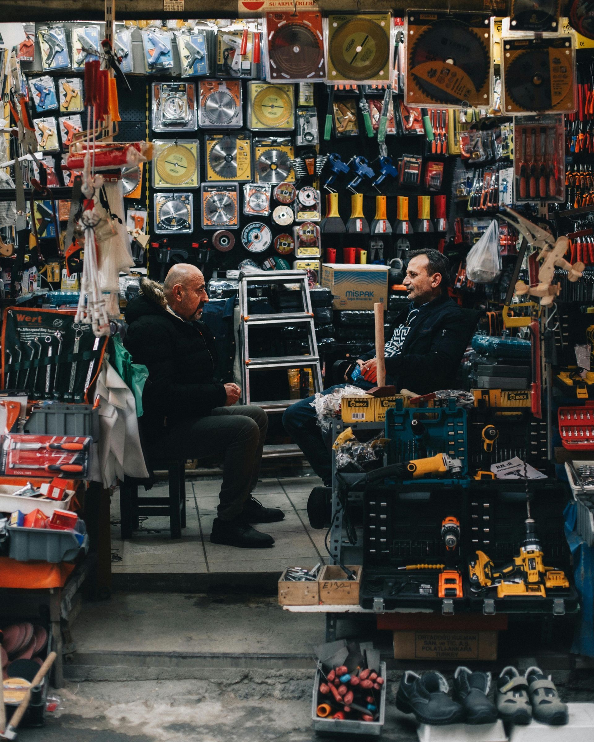 Two men conversing in a hardware store. Tools and supplies surround them. One holds an item overhead.