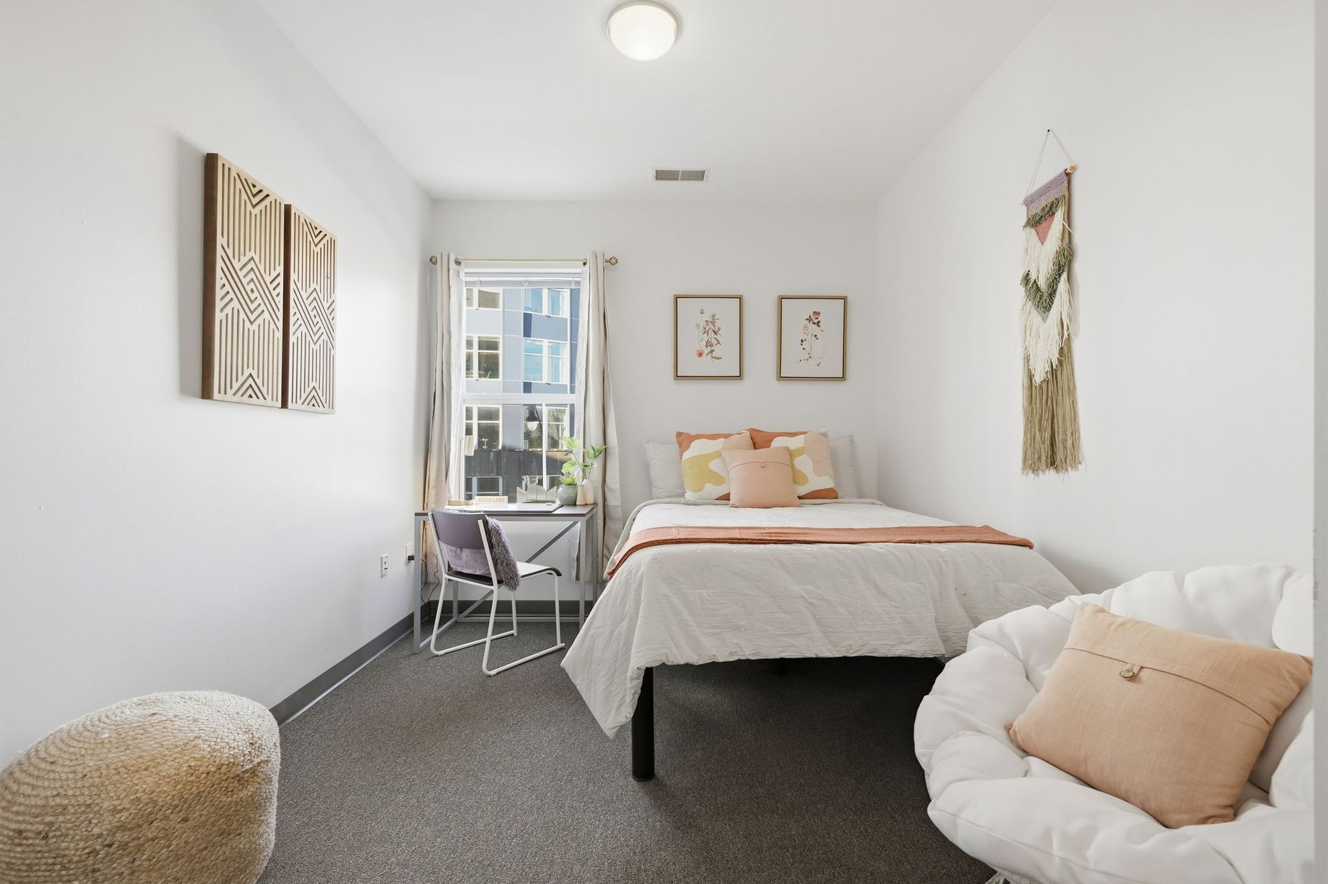 A small bedroom with a bed, desk, and chair. White walls, light-colored bedding, and a neutral color scheme.