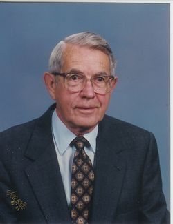 Older man wearing glasses, suit, and patterned tie, posing for a portrait against a blue backdrop.