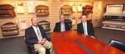 Three men in suits sitting at a table in a showroom with caskets displayed on the wall.