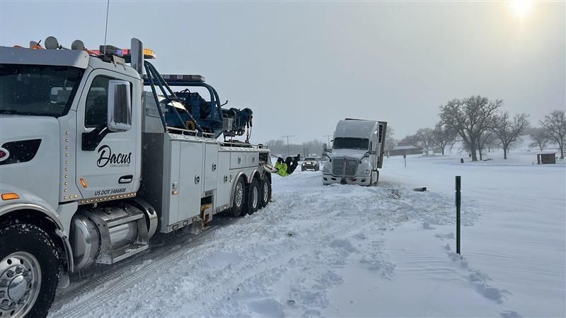 A blue tow truck lifting a white container from a teal semi-truck on a gravel lot under a blue sky.