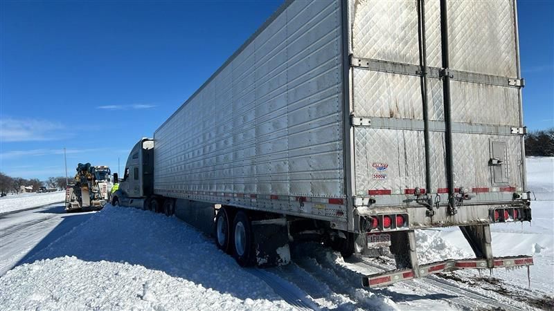 Semi-truck stuck in snow on the side of a road, tow truck in the distance, clear blue sky.