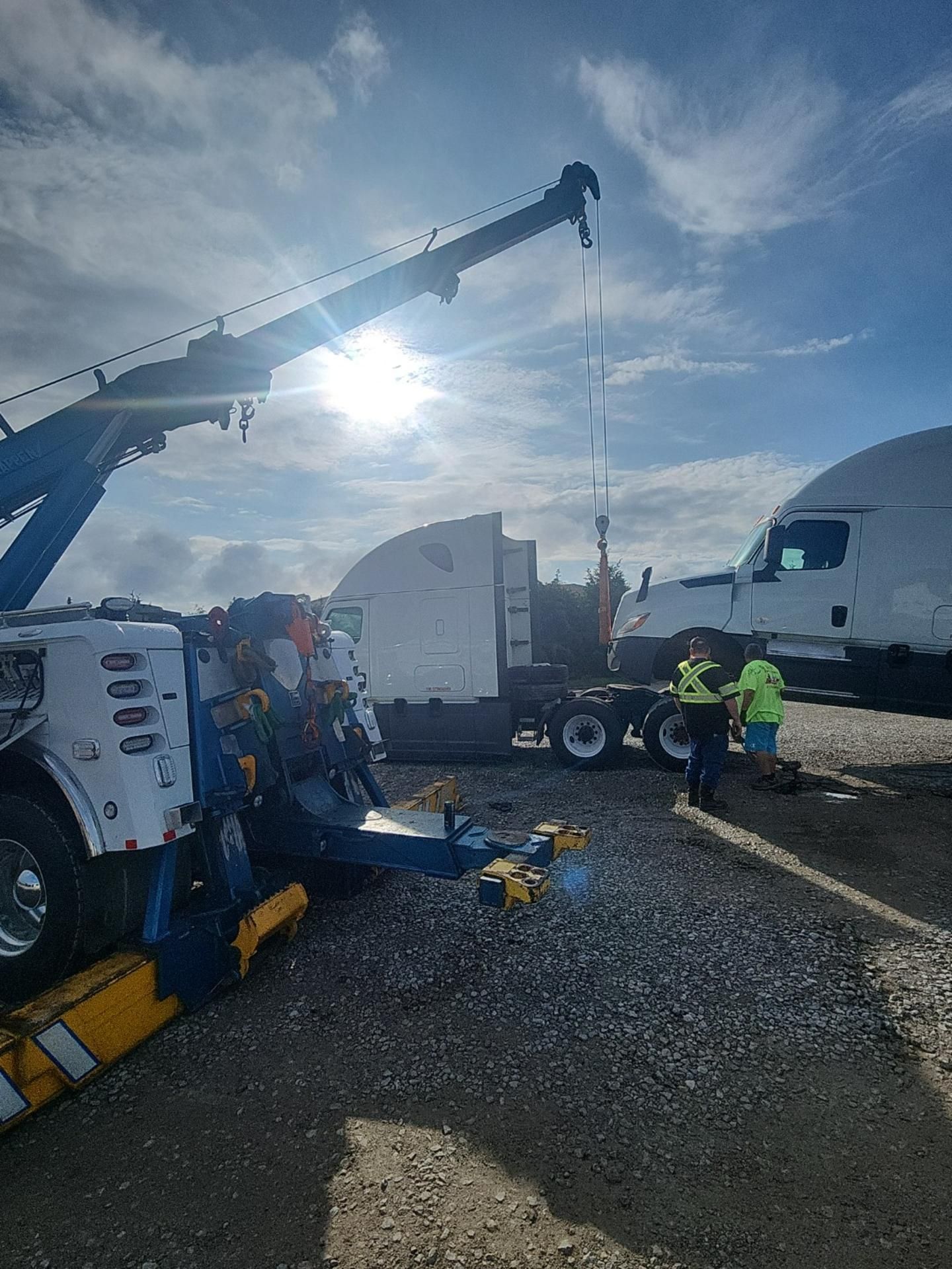 Tow truck lifting a semi-truck cab. Two people in safety vests assist. Bright sunlight, cloudy sky.