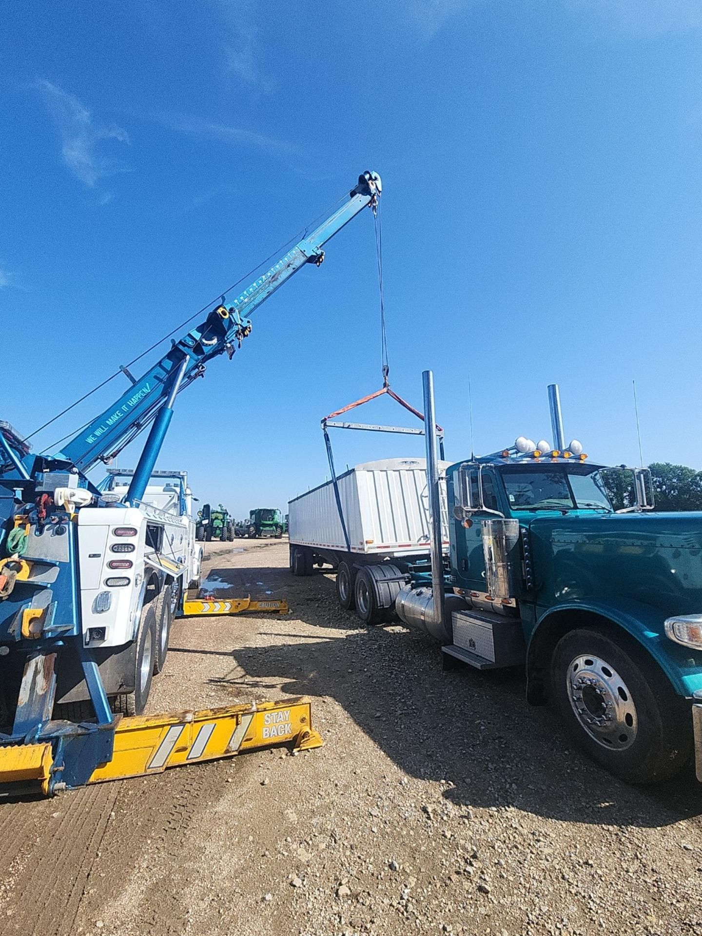 A blue tow truck lifting a white container from a teal semi-truck on a gravel lot under a blue sky.