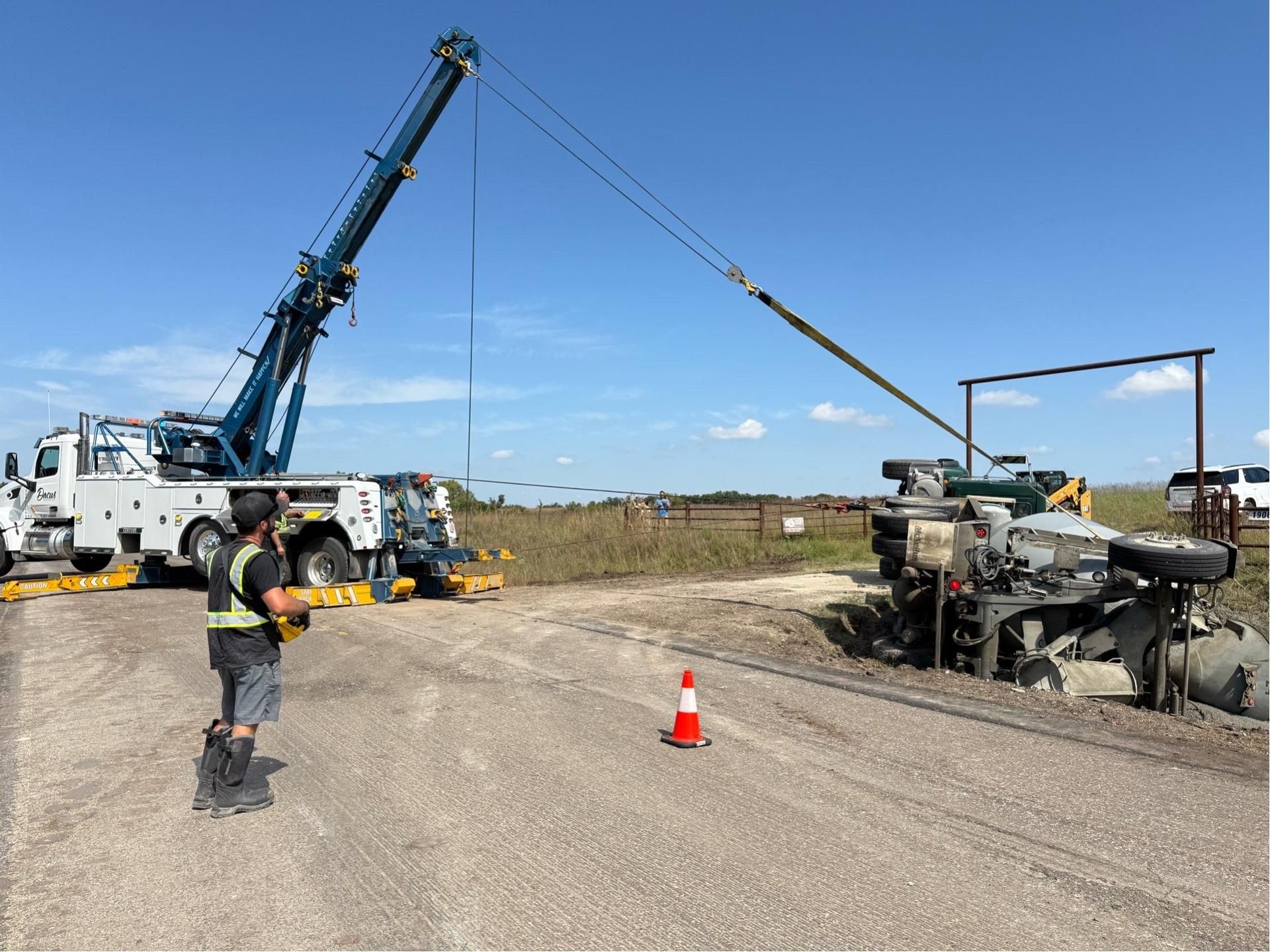 Crane lifting equipment next to a truck on a gravel road, two workers, clear sky.