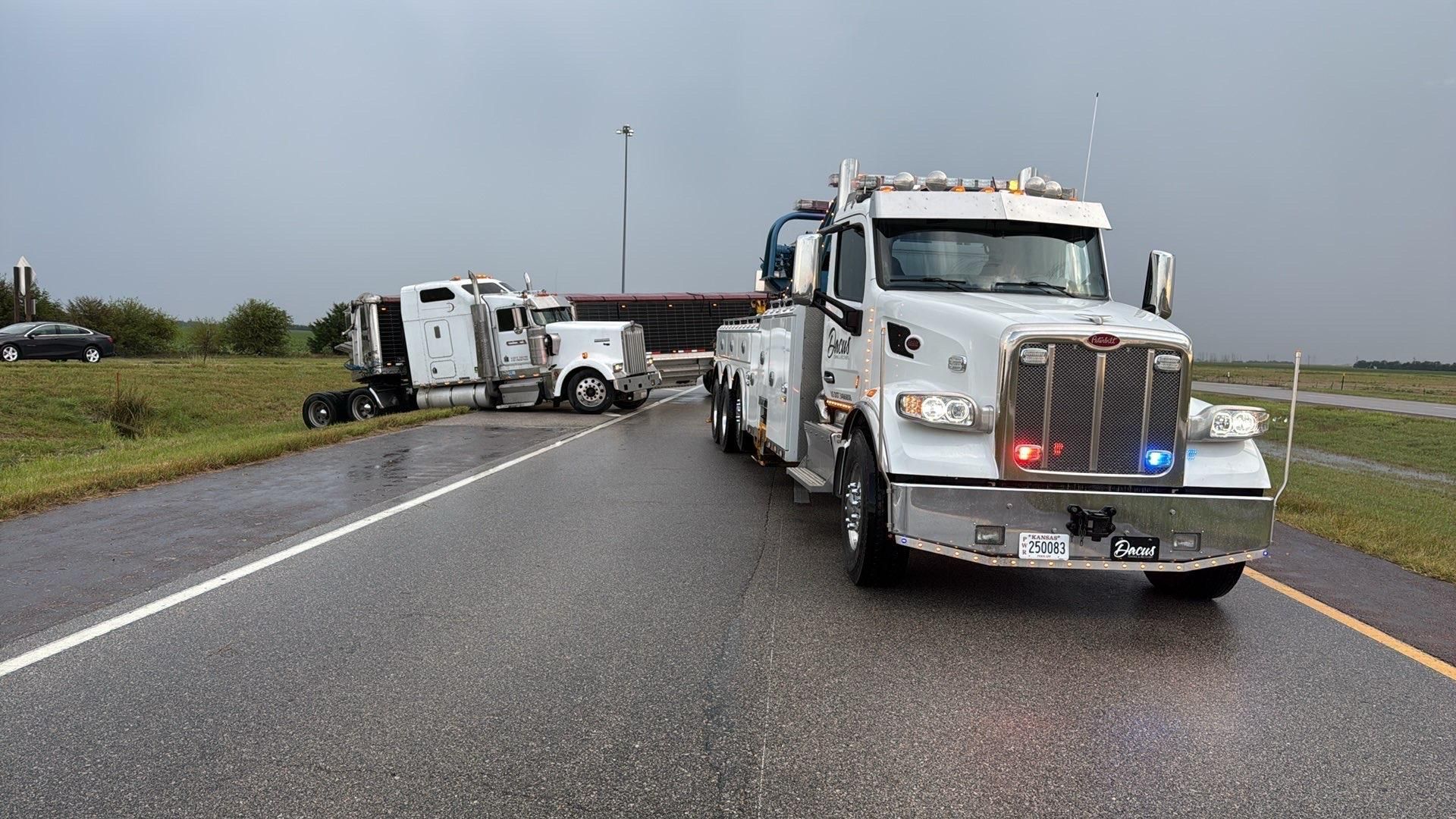 A tow truck on a wet highway scene with a tipped-over semi-truck under a bridge on the side.