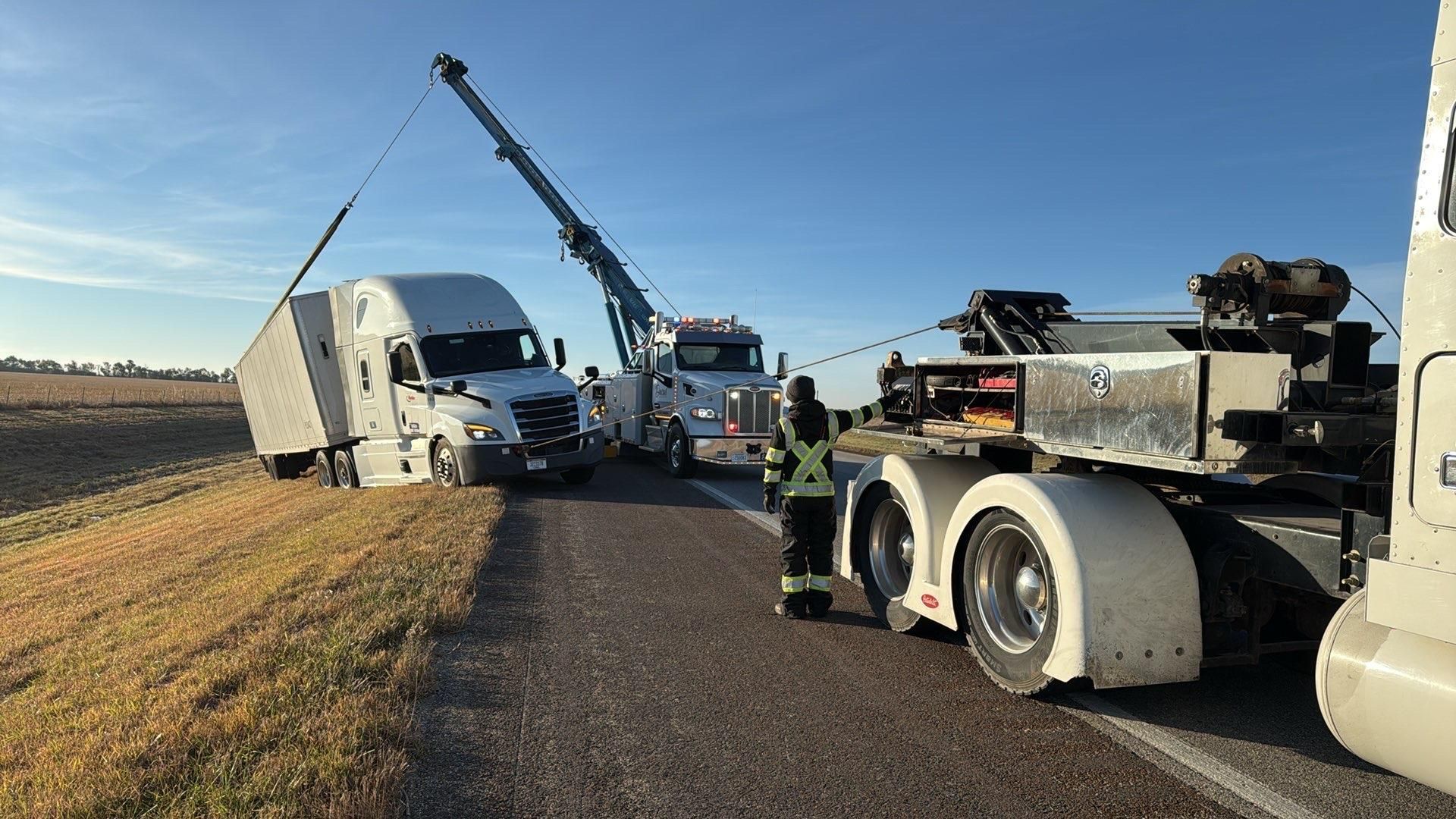 Tow truck lifting a semi-truck cab. Two people in safety vests assist. Bright sunlight, cloudy sky.
