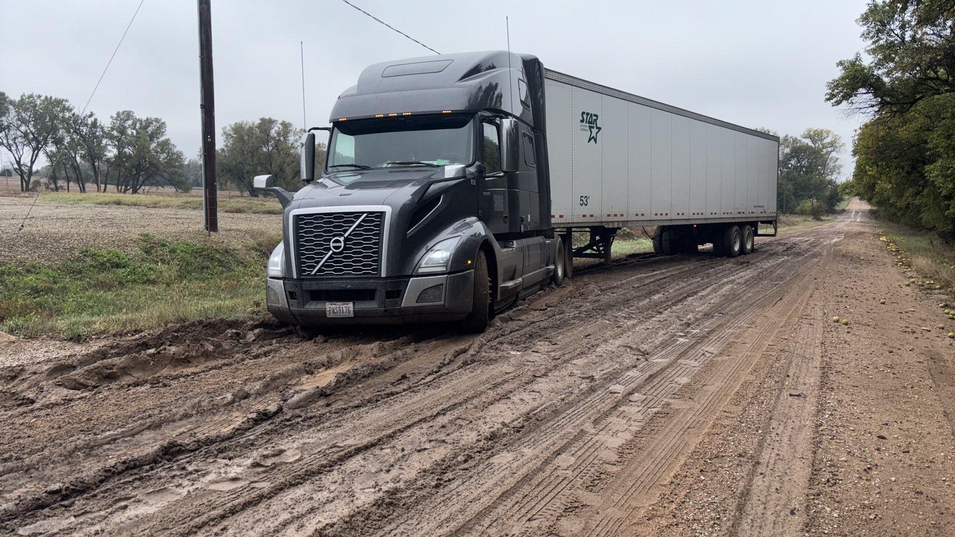 Semi-truck stuck in deep mud on a dirt road. Gray truck, white trailer. Overcast sky.