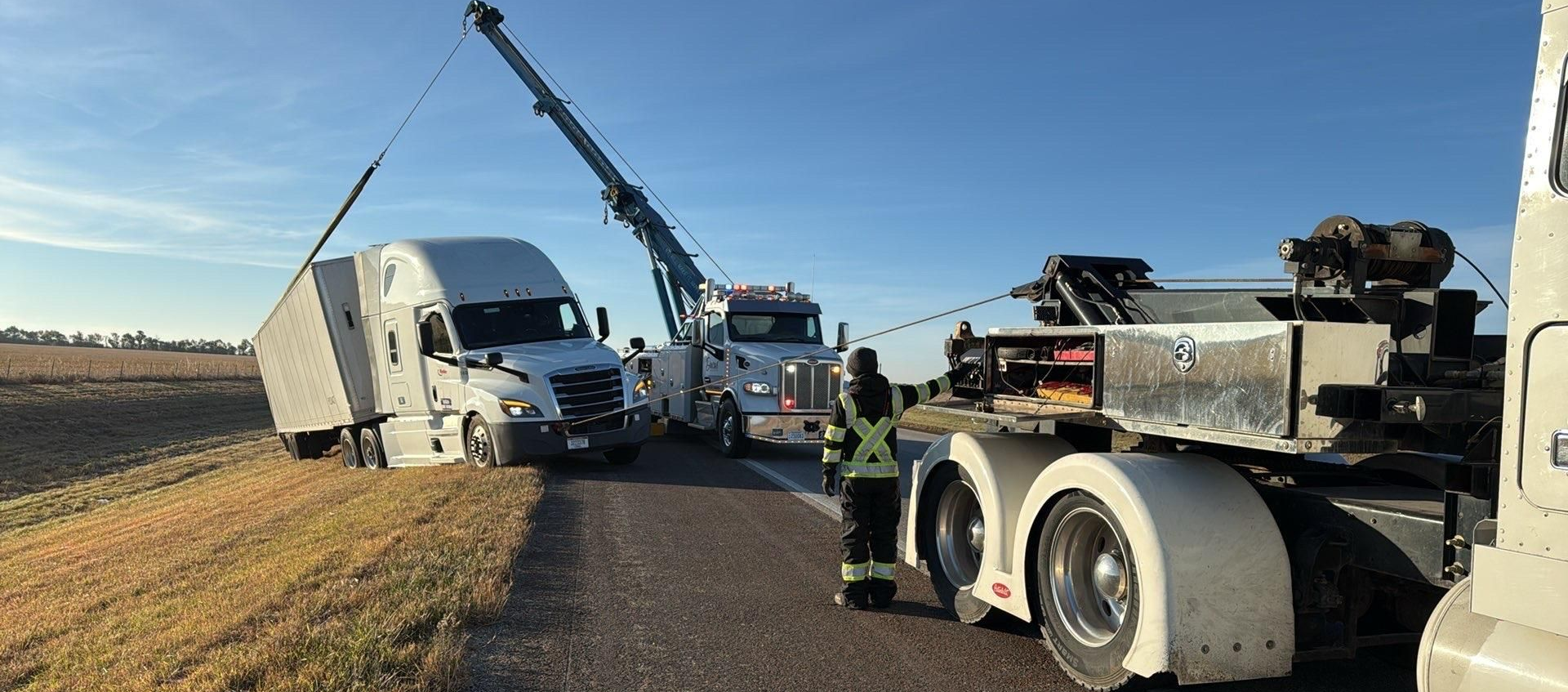 A semi-truck being towed from a ditch by a tow truck on a rural road under a blue sky.