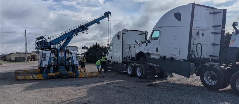Semi-truck stuck in snow on the side of a road, tow truck in the distance, clear blue sky.