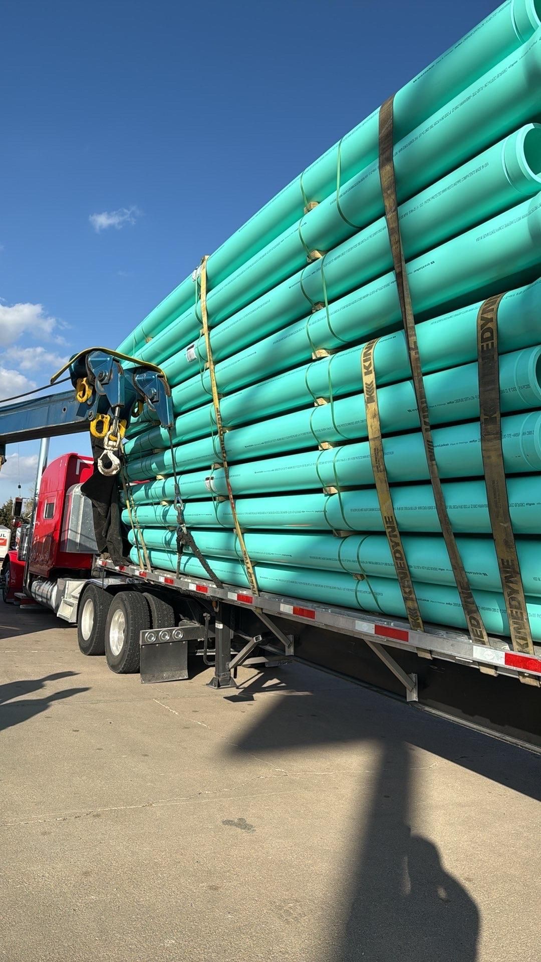 A semi-truck loaded with teal pipes secured by straps under a crane against a blue sky.