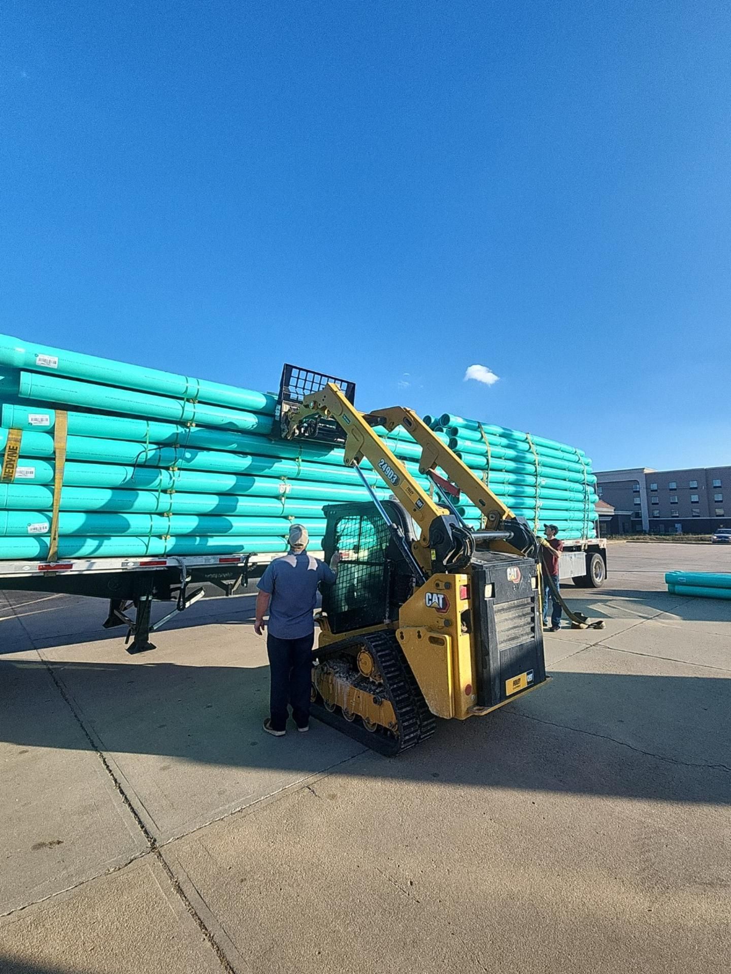 A yellow skid steer unloading green pipes from a trailer under a blue sky. Two workers observe.