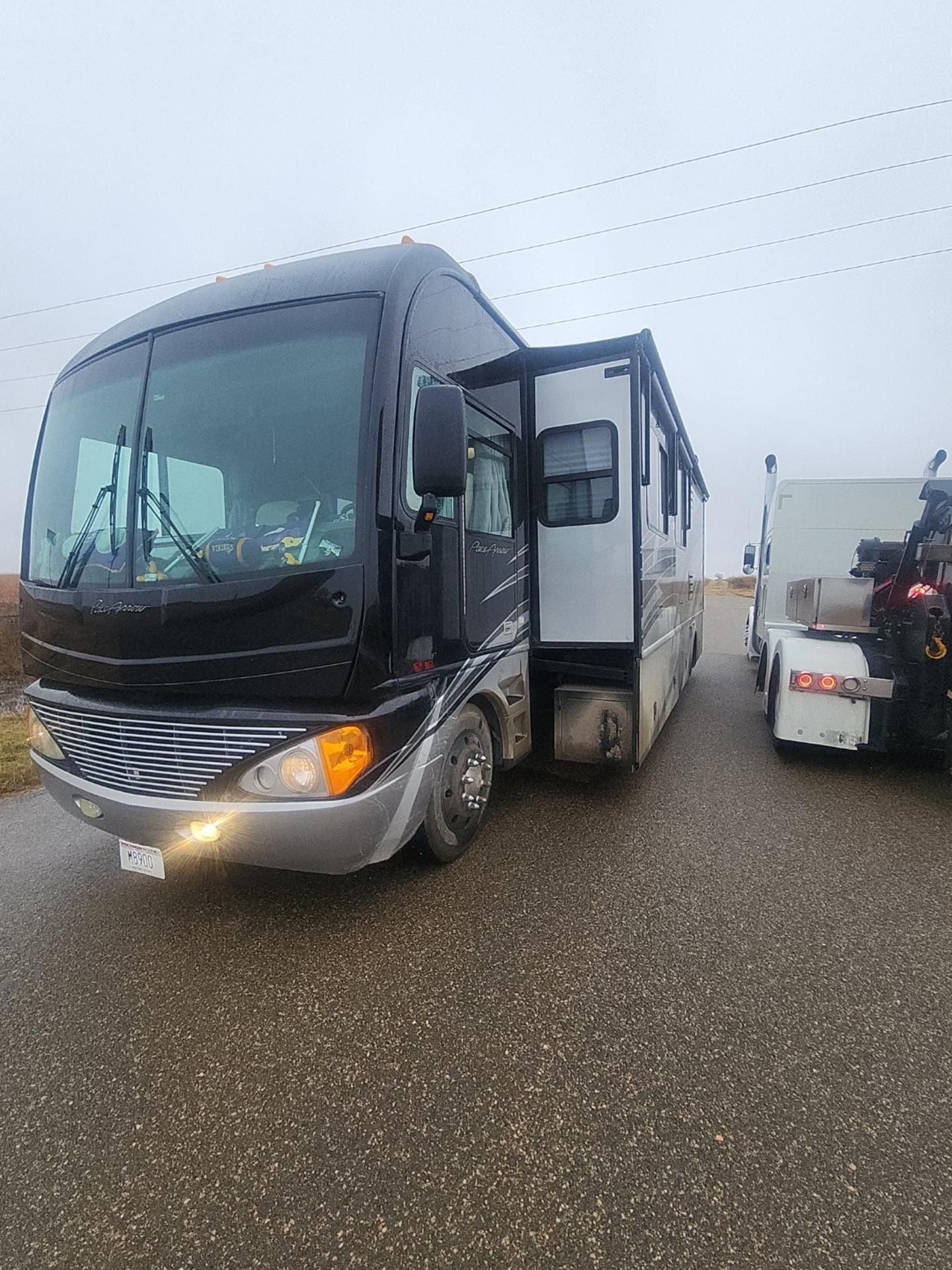 Black and silver RV with open door on a wet road, next to a white truck. Overcast sky.