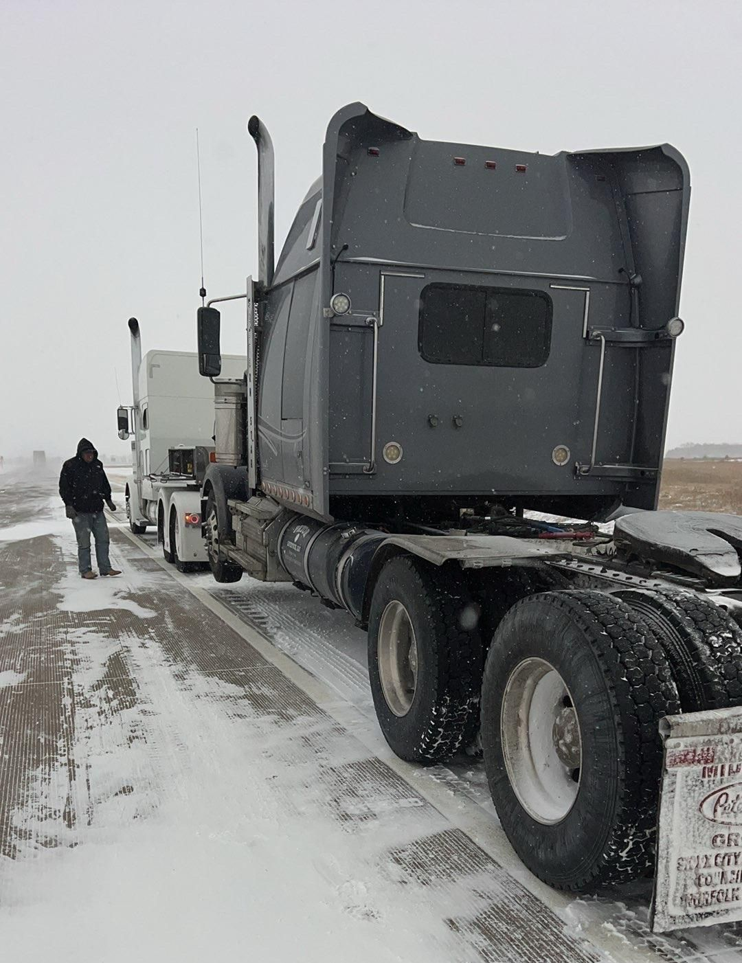 Two semi-trucks on snow-covered road. Person stands beside the white truck. Gray sky.
