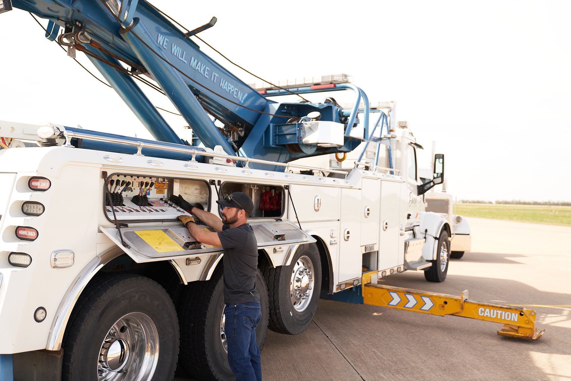 A tow truck operator working on the control panel of a white and blue tow truck, outdoors.