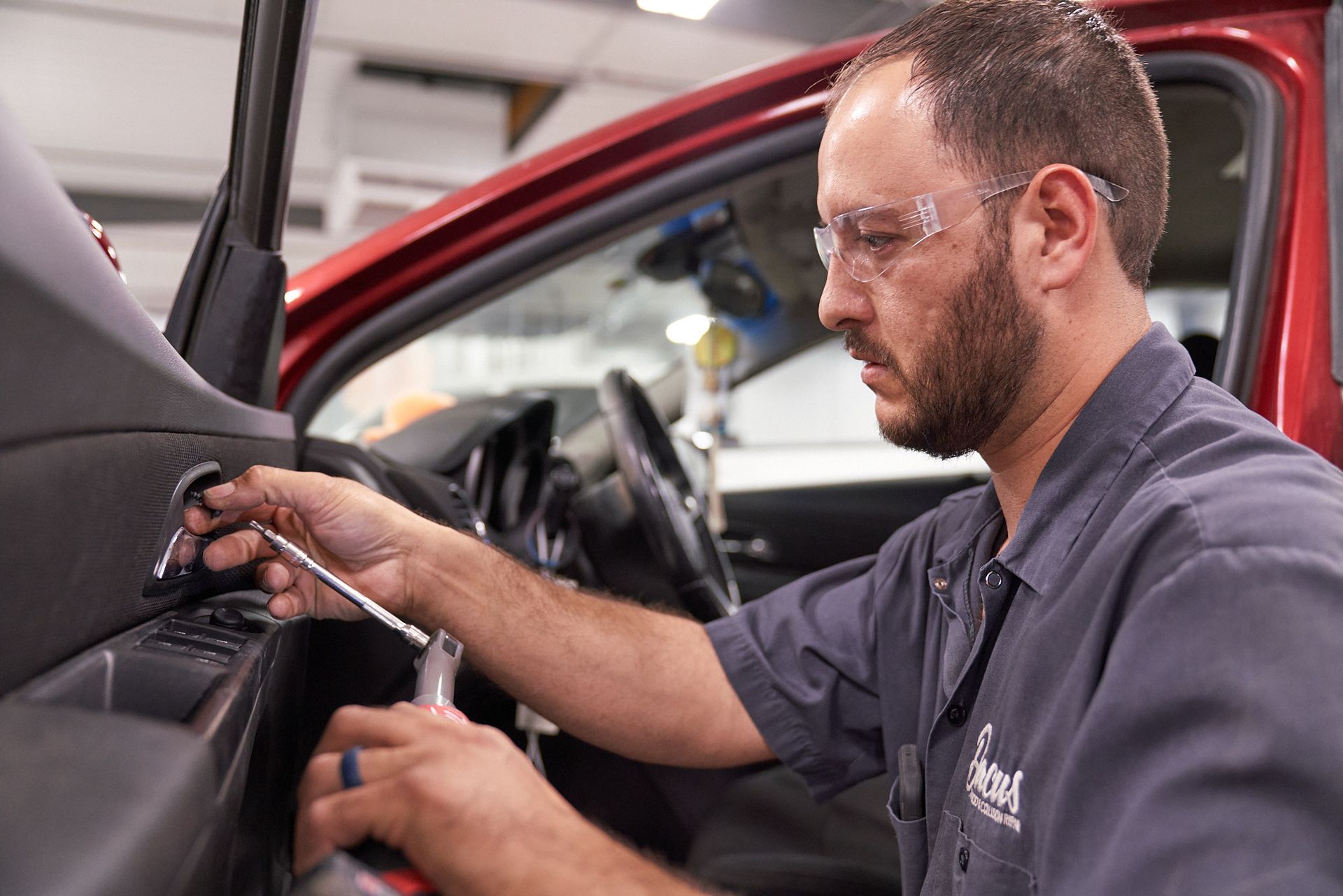 Mechanic works on a car door, wearing safety glasses. Red car, gray shirt, shop setting.