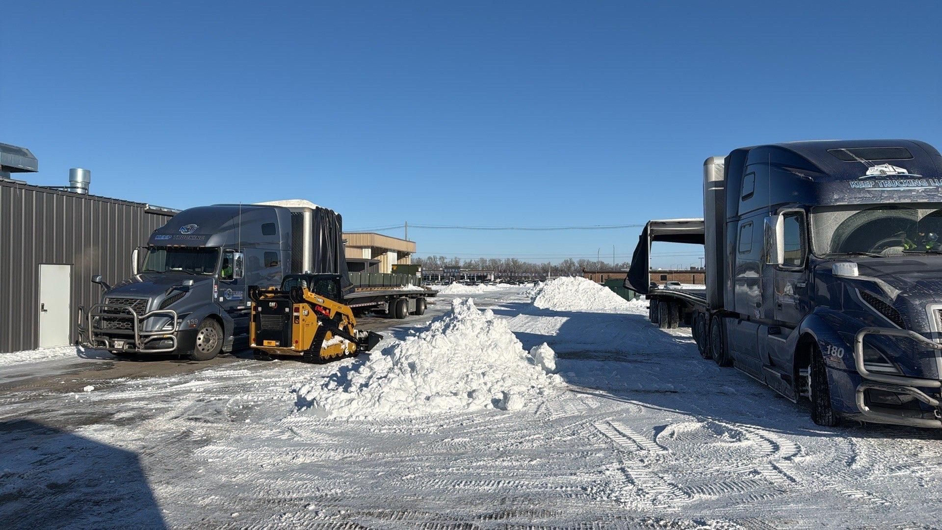 Two semi-trucks, one with a snowplow, clear a snowy area.