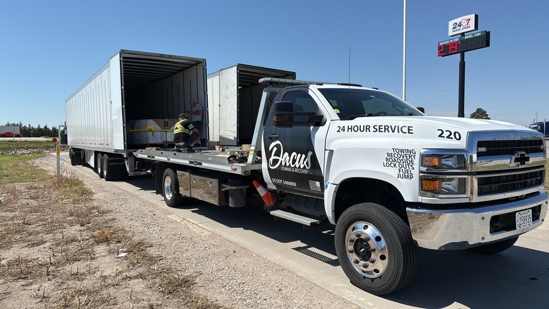 White tow truck hauling a semi-trailer on a roadside under a sunny sky.