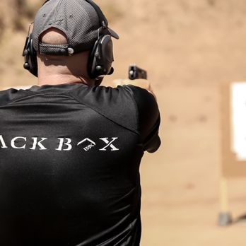 A man in a black shirt and cap shoots a handgun at a target outdoors; a shooting range.