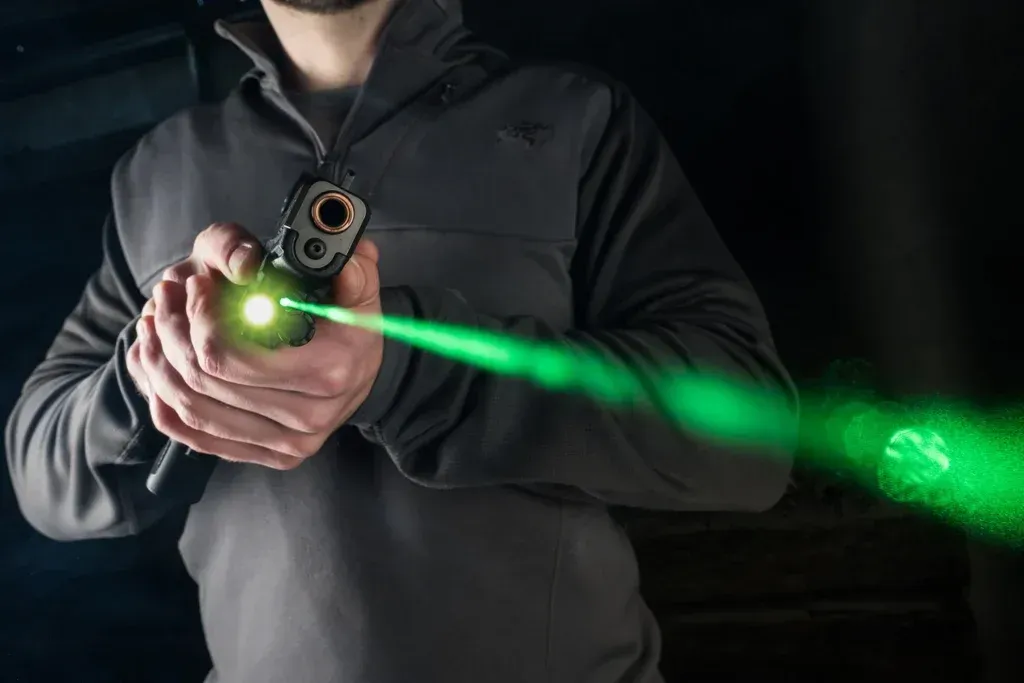 Person holding a black handgun with a bright green laser beam in a dark setting.