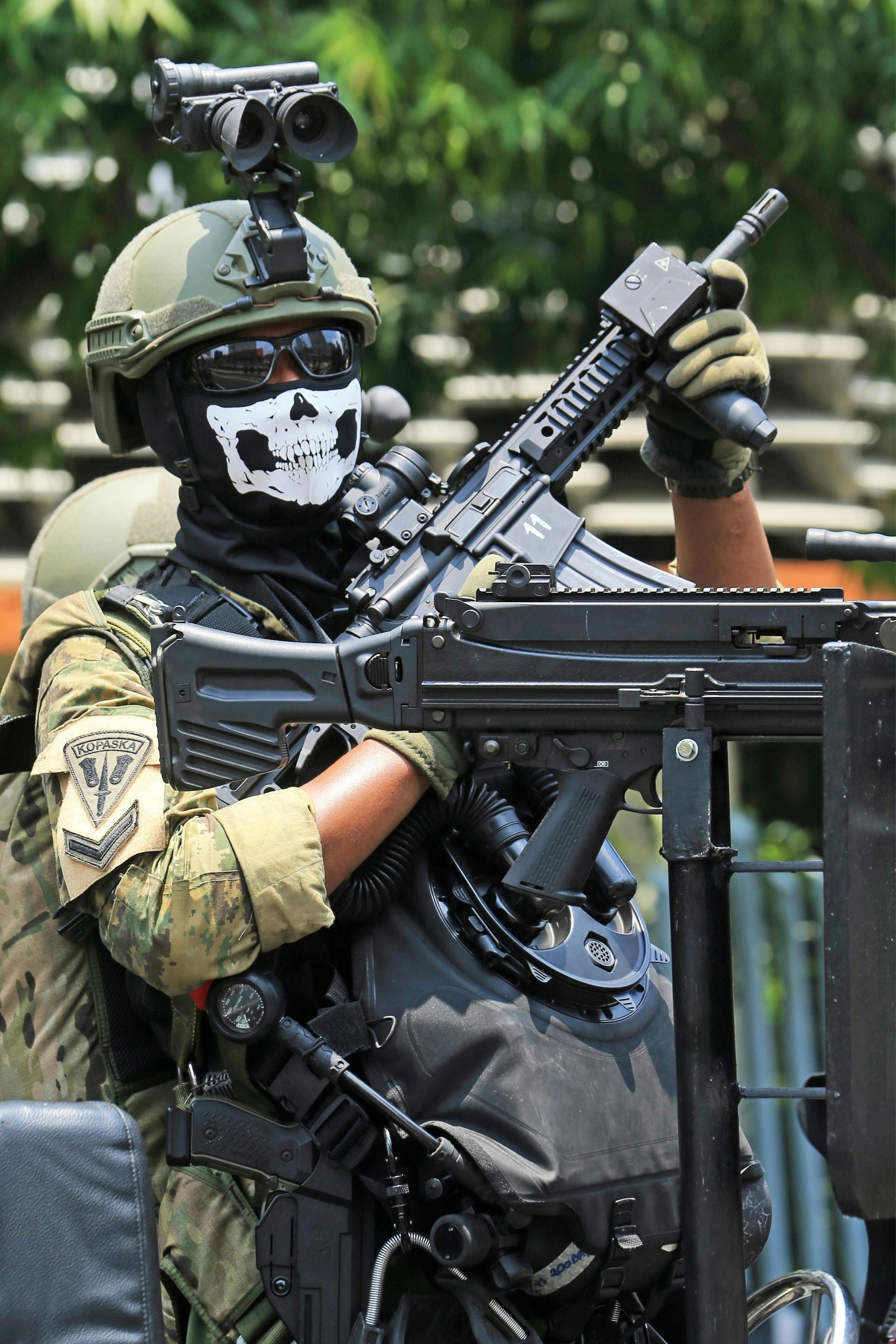 Soldier in camouflage, skull mask, and helmet holding a gun, sitting outdoors.