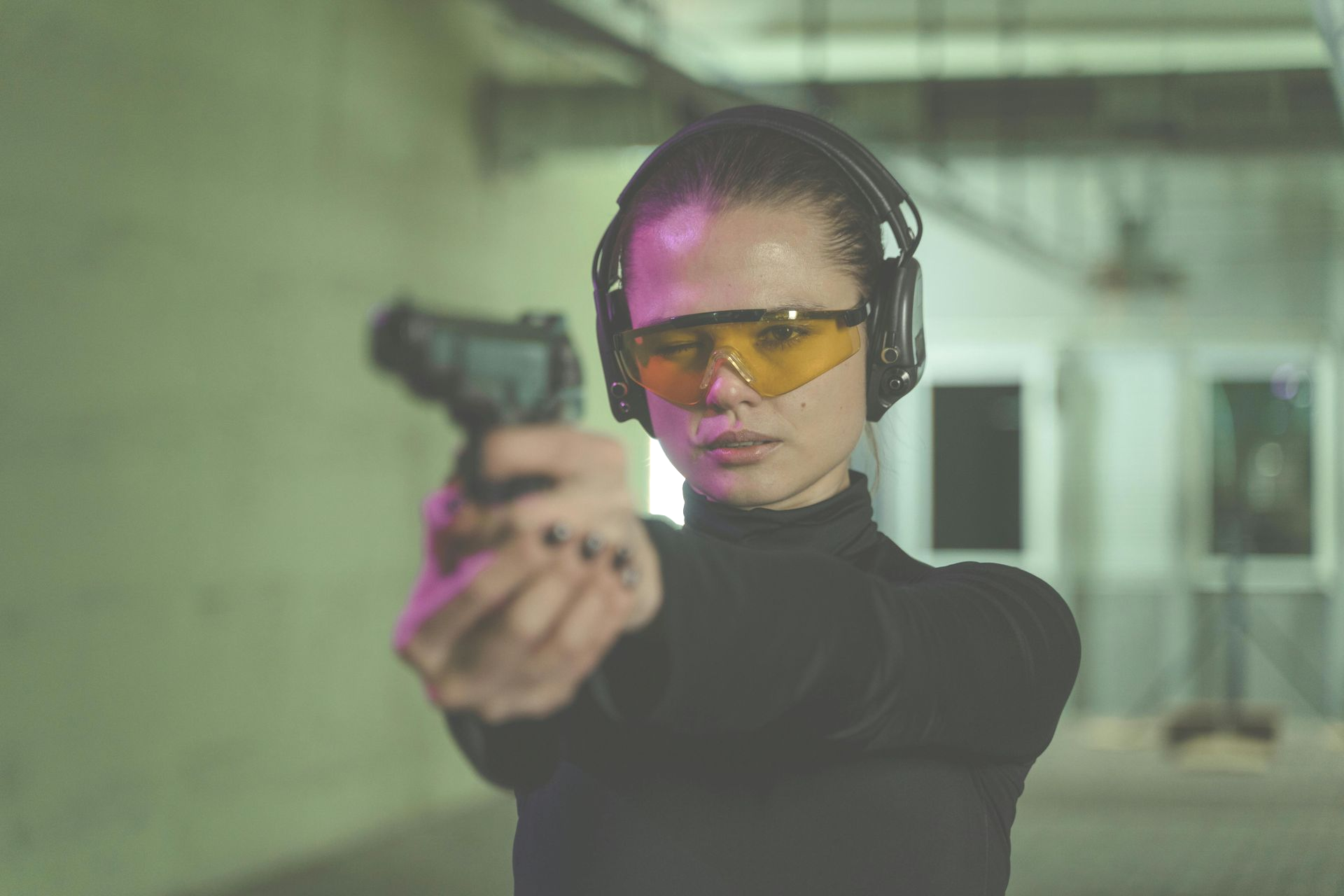 Woman in safety gear, holding a handgun at a shooting range, aiming at the target.