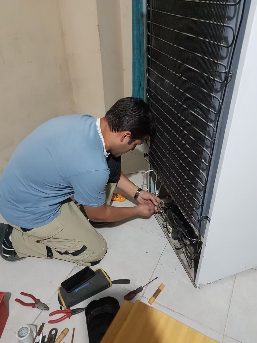 Hombre arrodillado reparando el compresor del refrigerador, con herramientas en el suelo. Interior, paredes neutras.