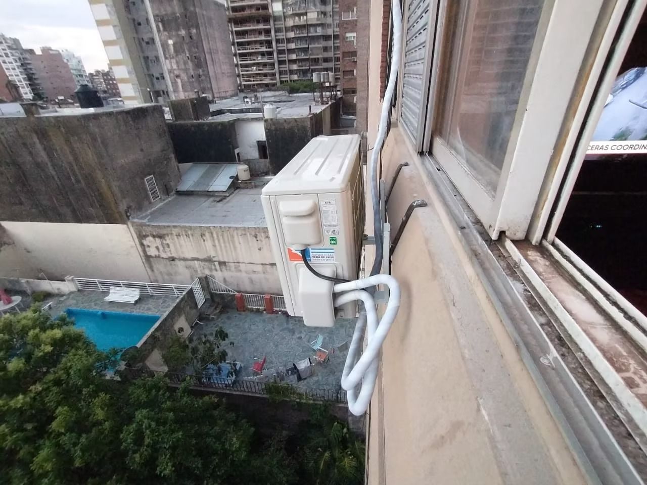 Unidad de aire acondicionado exterior montada en la pared de un edificio, con tuberías de conexión y tejados cercanos.