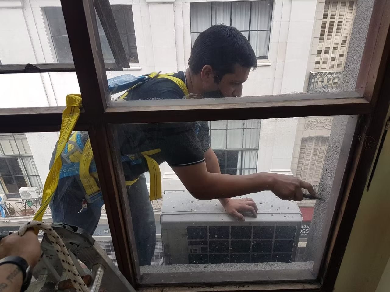 Hombre limpiando una ventana con un raspador. Lleva arnés y está cerca de un aparato de aire acondicionado.