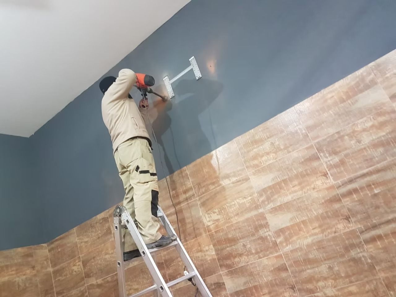 Una persona en una escalera instala algo blanco en una pared azul. Debajo, una pared de azulejos marrones.