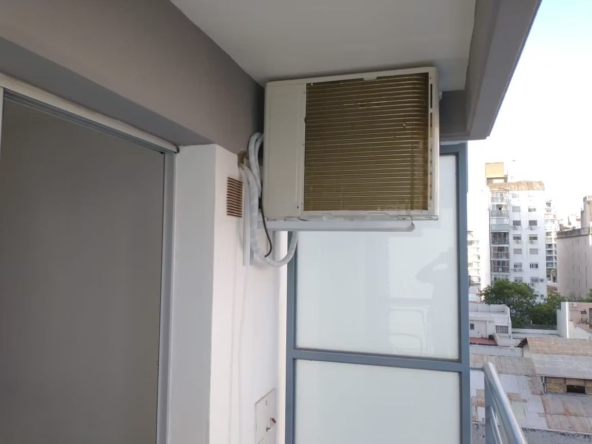 Unidad de aire acondicionado montada en una pared blanca, en un balcón con vista a los edificios.