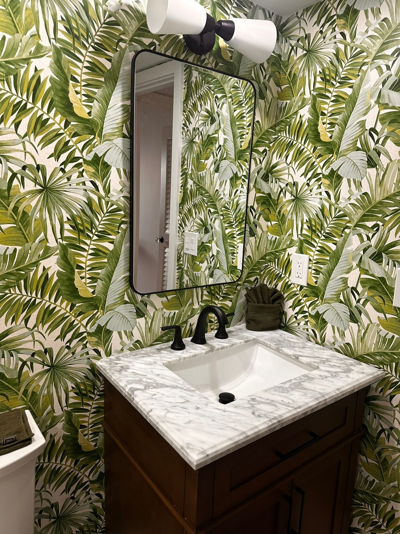 A bathroom with a sink , mirror , and tropical wallpaper.