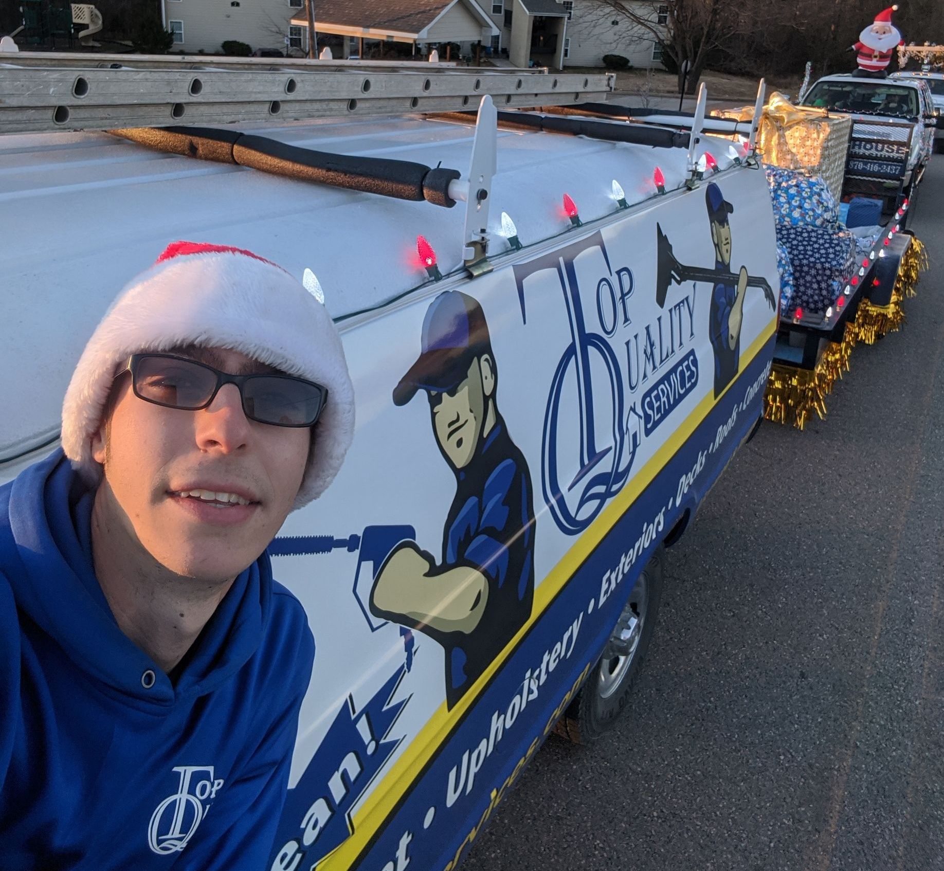A man is wearing a red Christmas cone hat standing in front of Top Quality Home Services Van in a Christmas parade line-up.