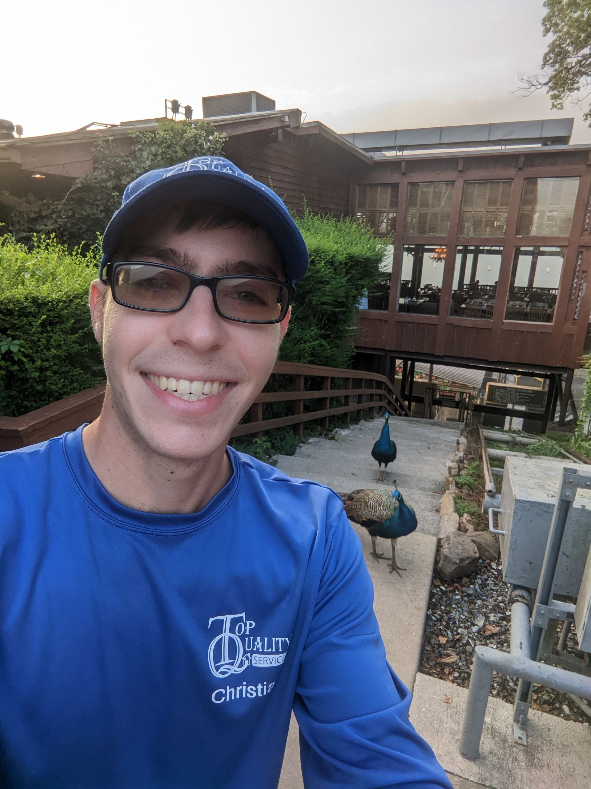 A man in a blue shirt is taking a selfie with a peacock in the background.