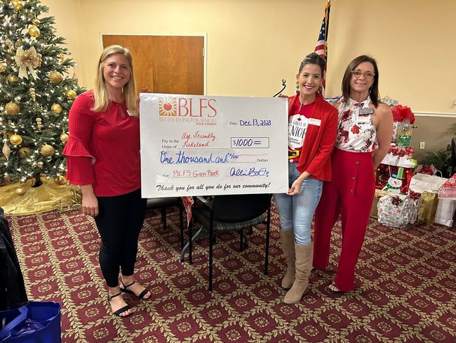 Three women are standing next to each other holding a large check in front of a christmas tree.