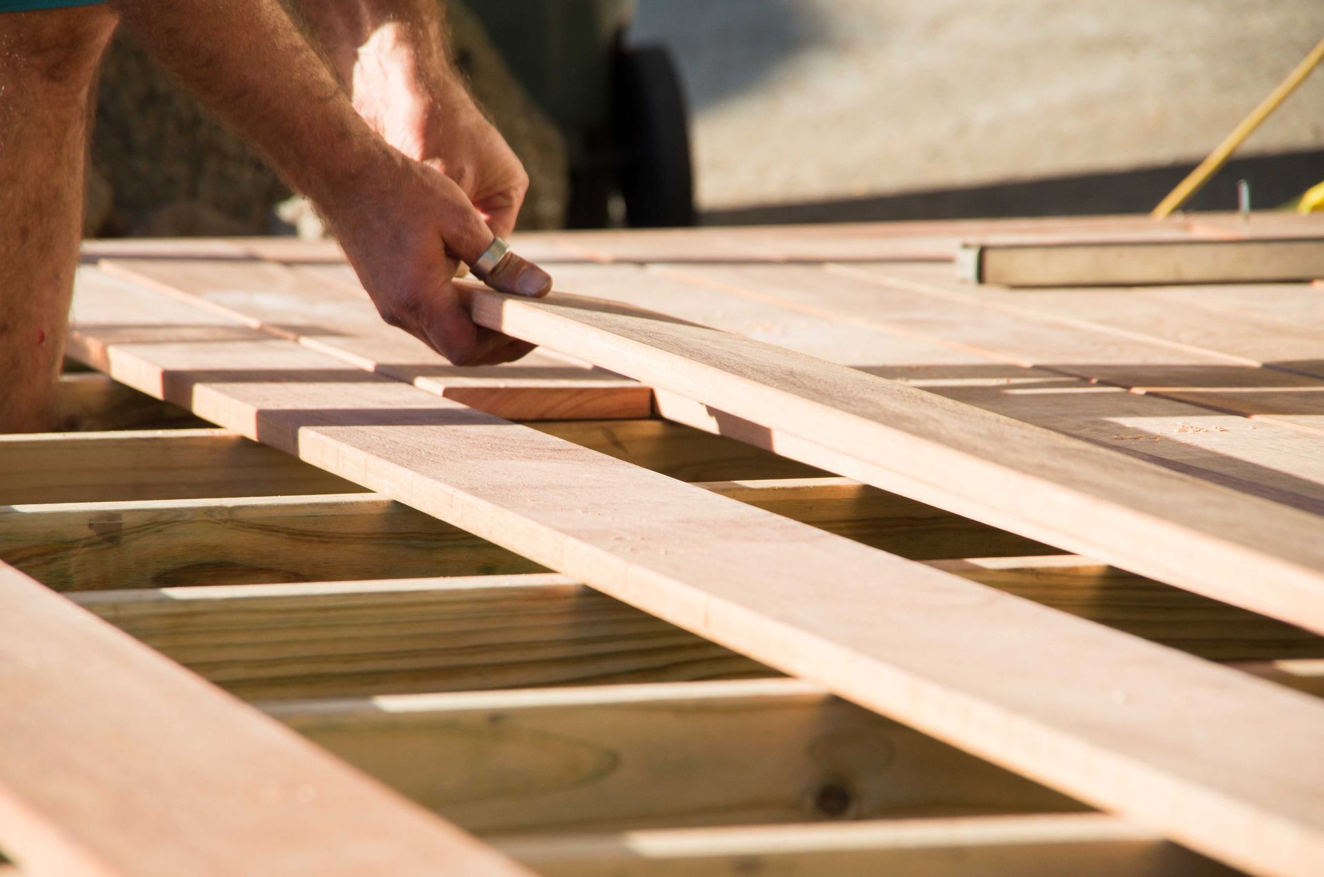 Une personne installe des planches de bois clair sur la structure d'une terrasse sous un éclairage ensoleillé.