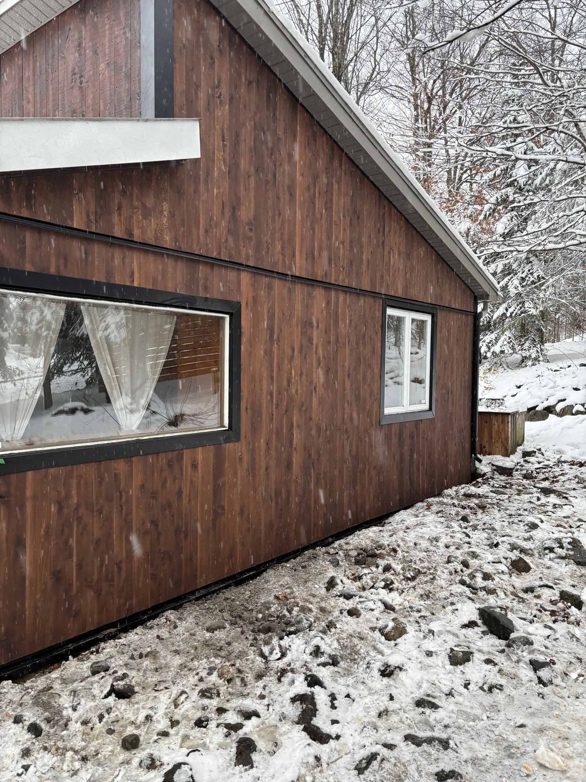 Vue latérale d'une cabane en bois brun avec deux fenêtres, située dans un paysage rocheux enneigé en hiver.