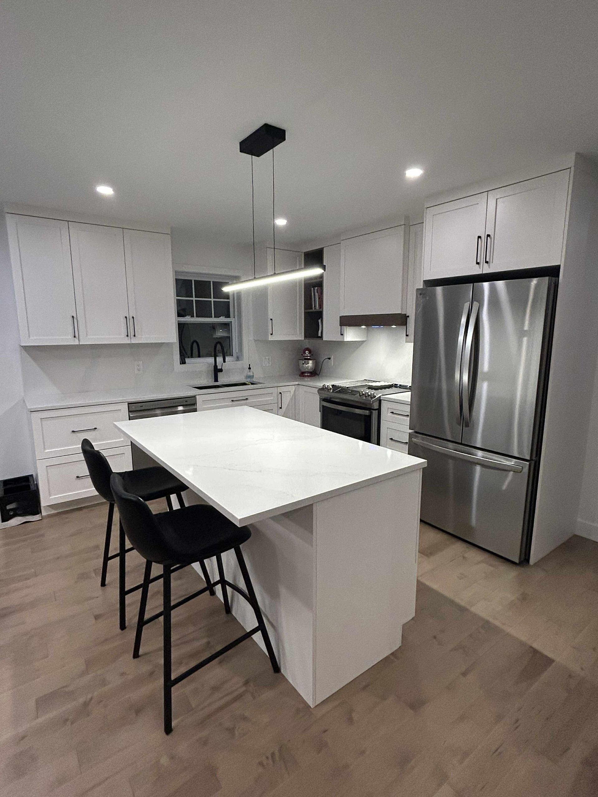 Une cuisine blanche moderne et minimaliste avec un îlot central, deux tabourets noirs, des appareils électroménagers en acier inoxydable et un sol en bois.