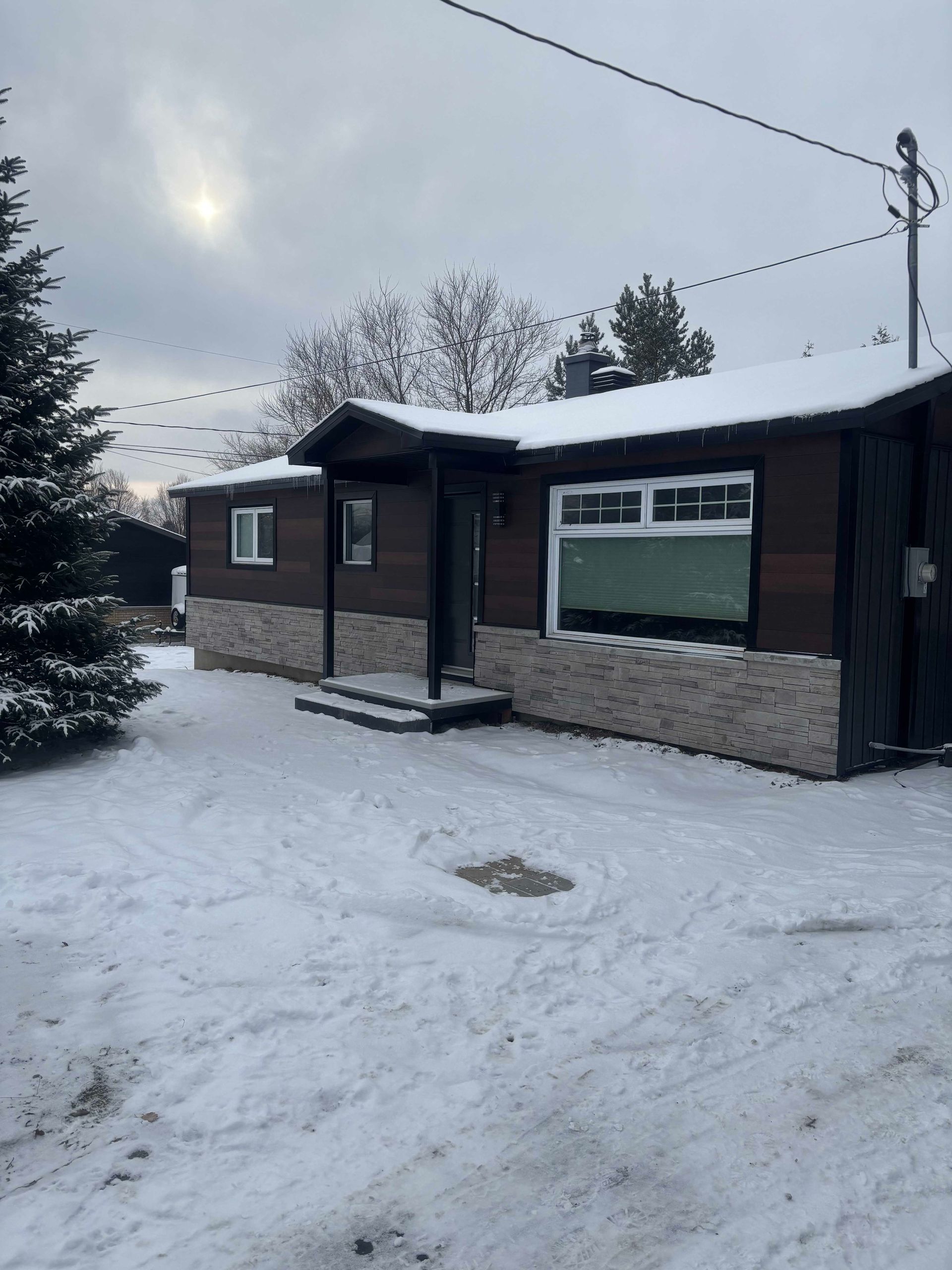 Une maison brun foncé, aux murs de pierre et au toit enneigé, située dans une cour recouverte de neige avec un conifère.