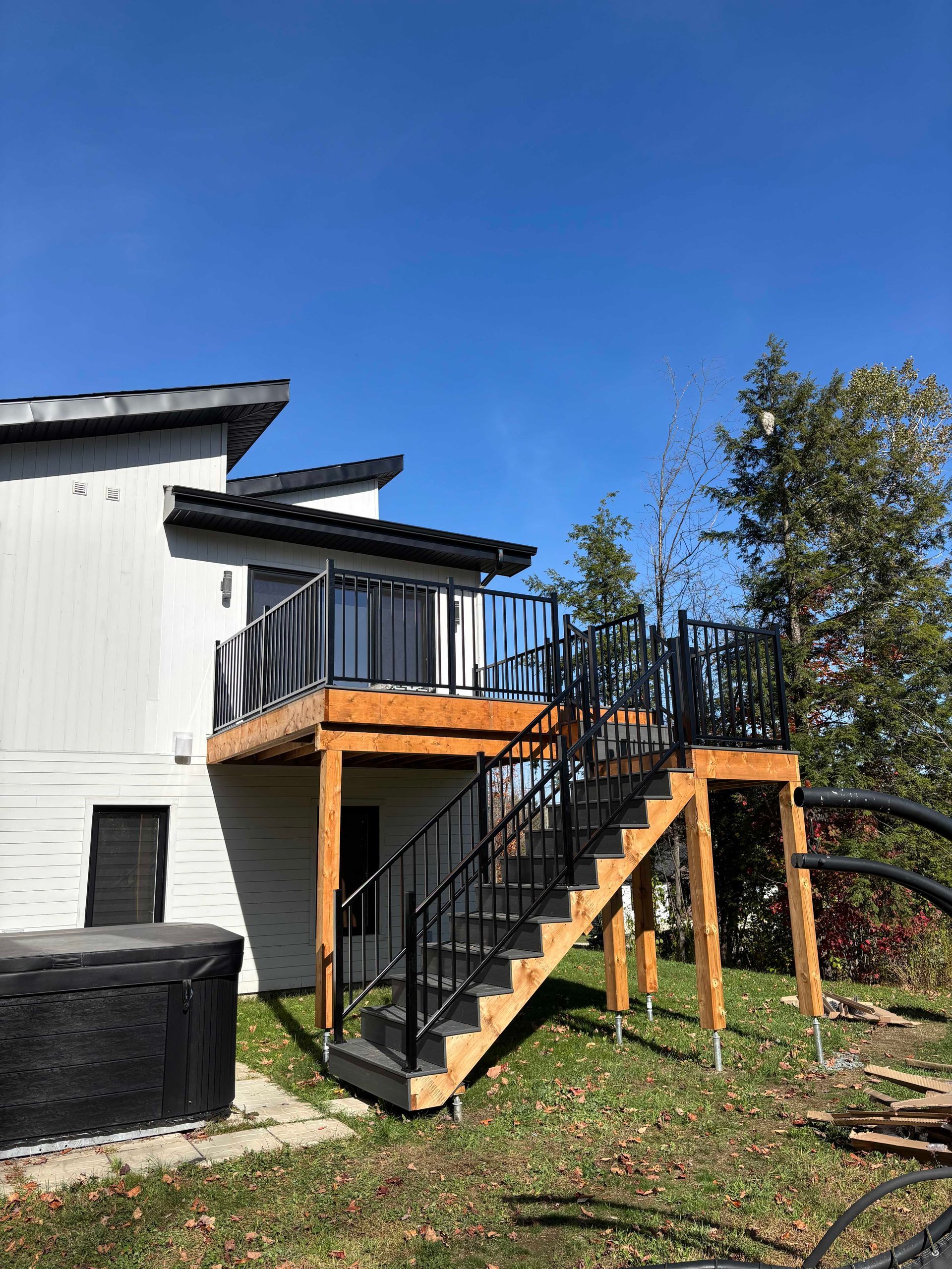 Vue extérieure d'une nouvelle terrasse en bois et d'un escalier en métal noir adossé à une maison, sous un ciel bleu dégagé.