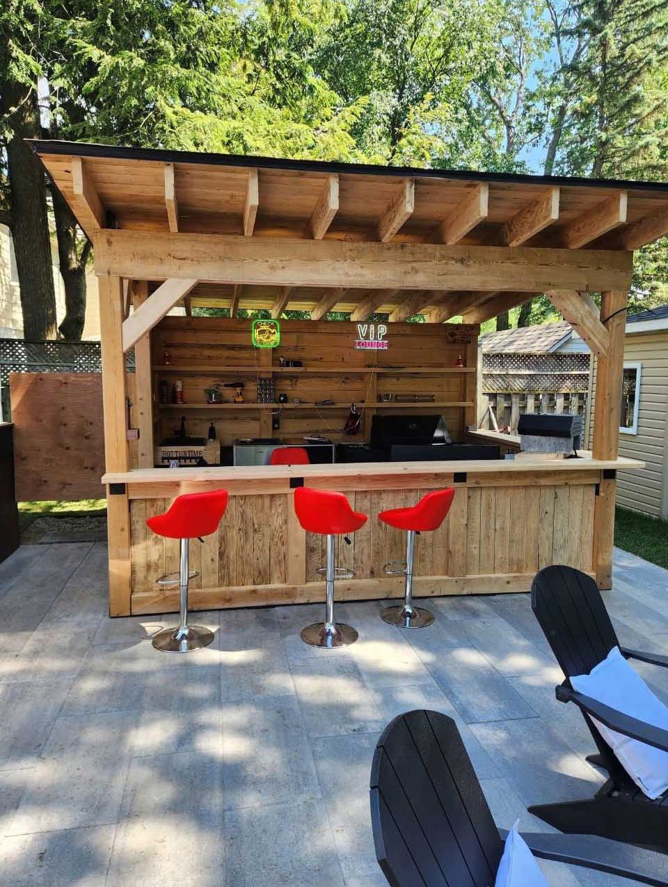 Une structure de bar extérieure en bois avec un toit, trois tabourets de bar rouges et des étagères sur une terrasse pavée dans une cour arrière.