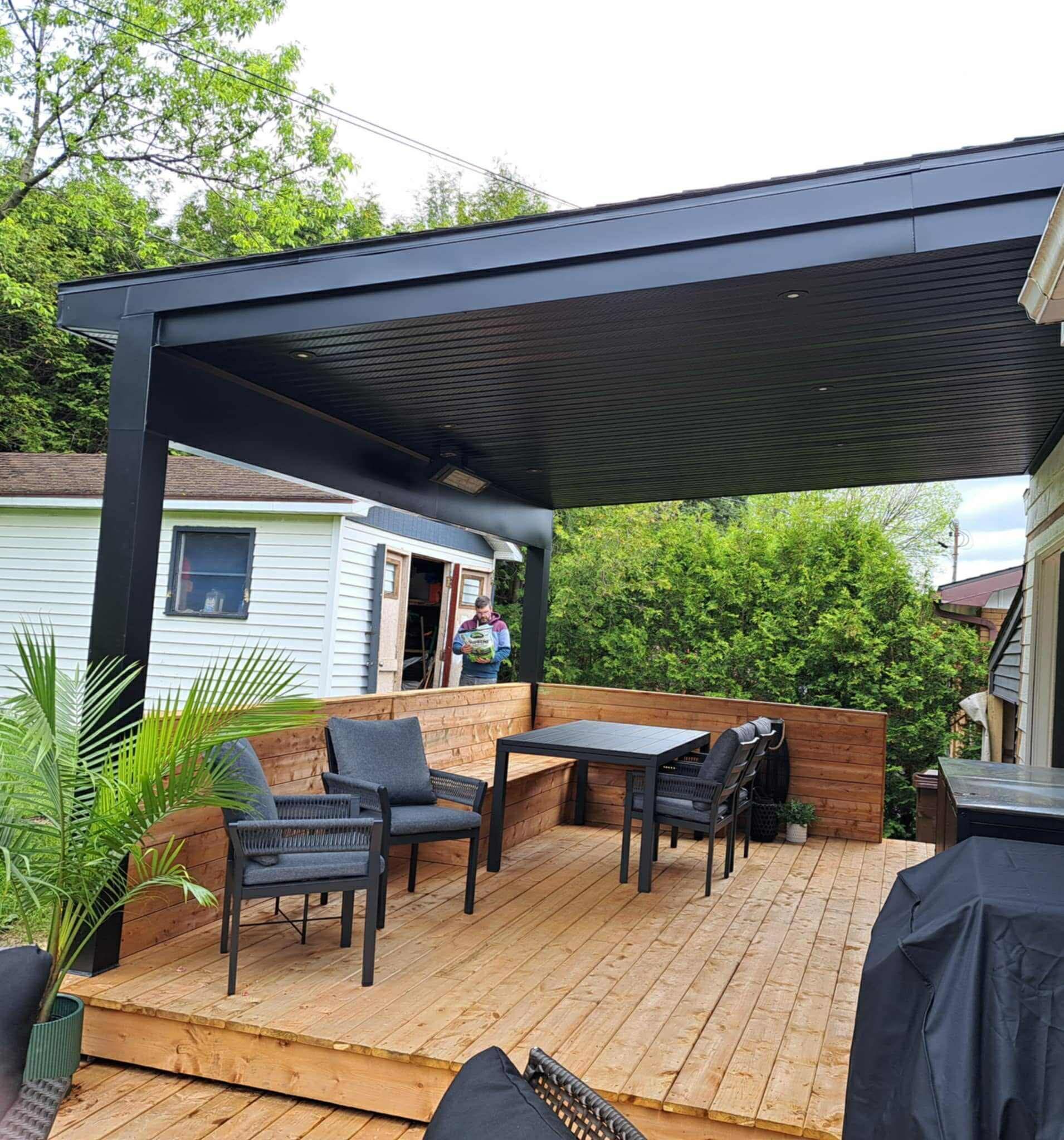 Une terrasse en bois avec une pergola noire abritant une table à manger, des chaises et un banc intégré dans une cour arrière résidentielle.