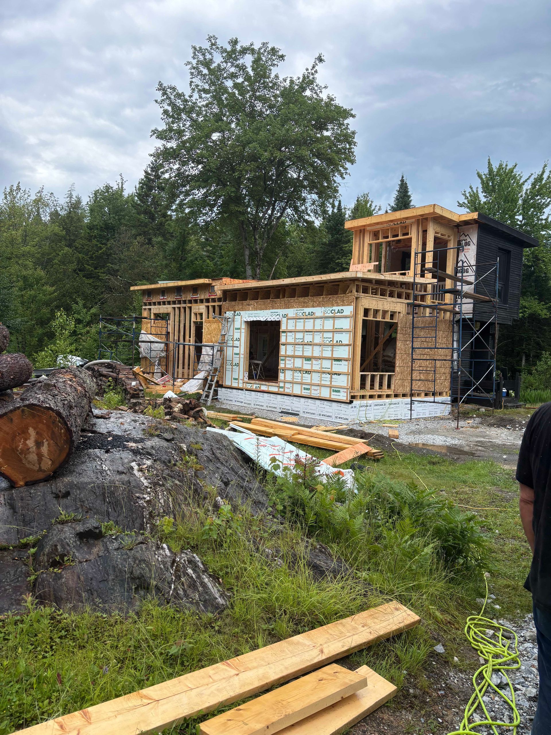 Une maison à ossature bois en construction se dresse dans un paysage rural et rocailleux, avec des arbres verdoyants sous un ciel nuageux.