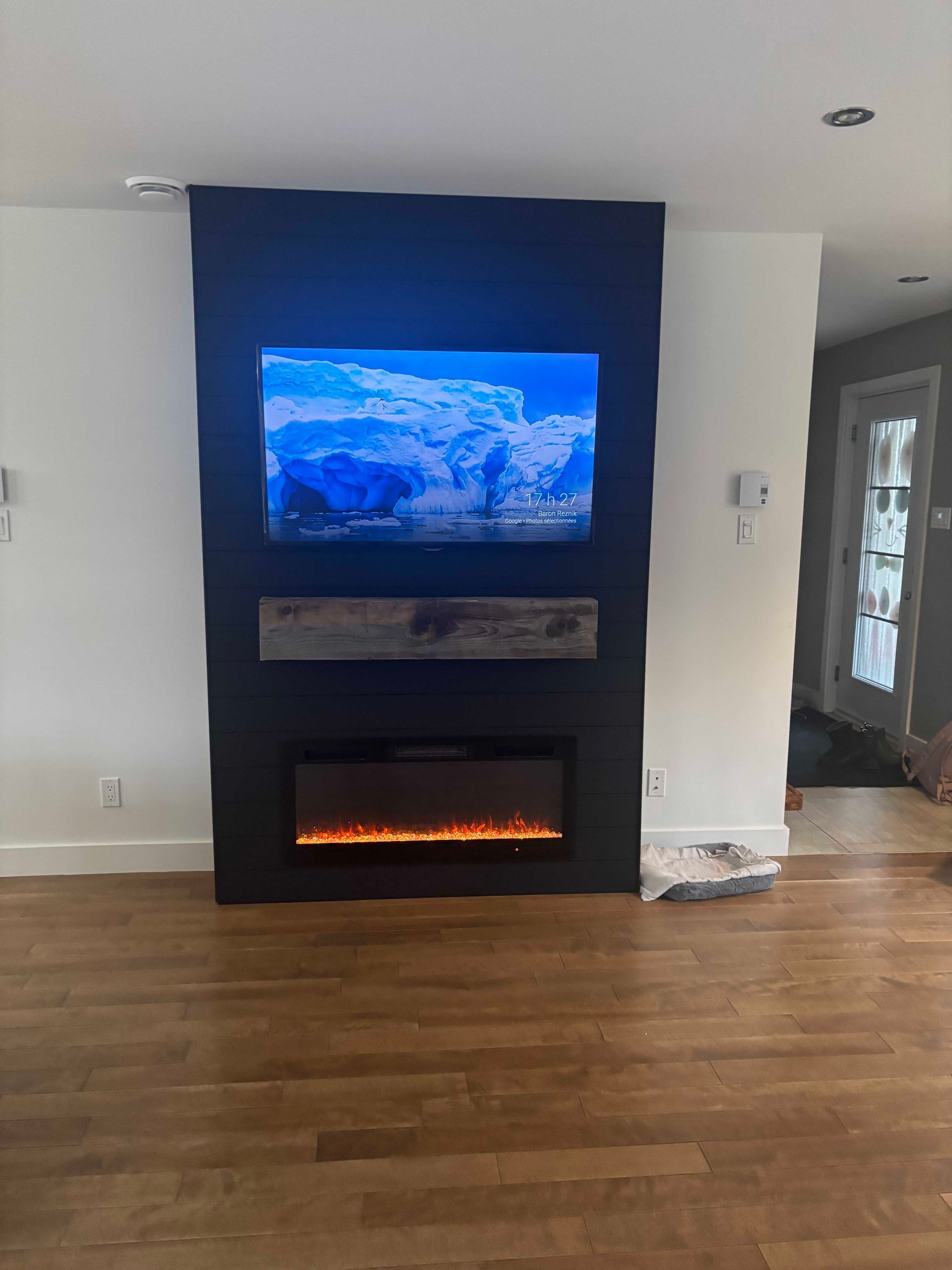 Dans un salon moderne et lumineux, une télévision murale et une cheminée électrique sont encastrées dans un mur rectangulaire sombre.