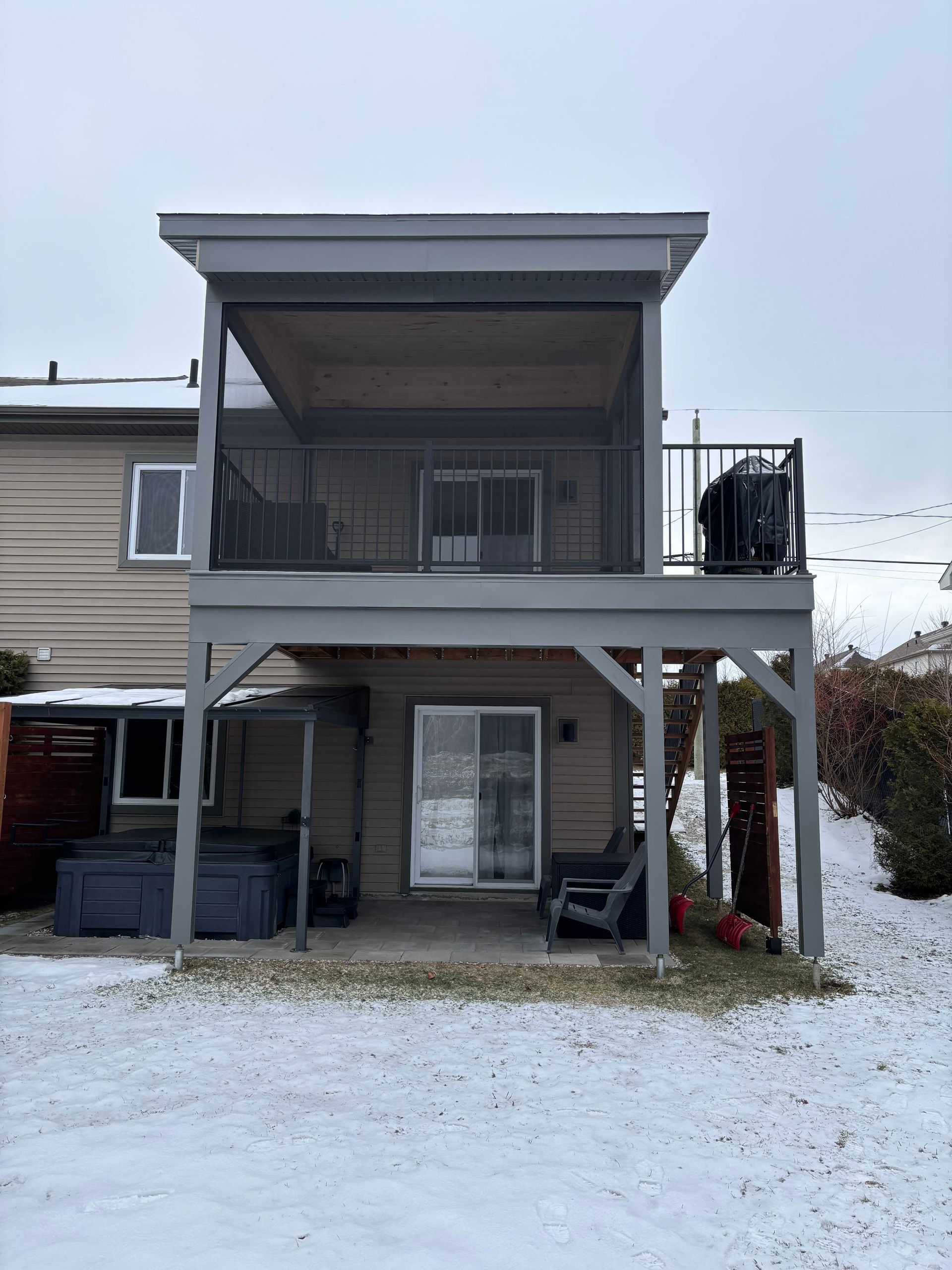 L'extérieur d'une maison à deux étages en hiver, avec une grande terrasse supérieure couverte, un patio en dessous et un jacuzzi sur la gauche.