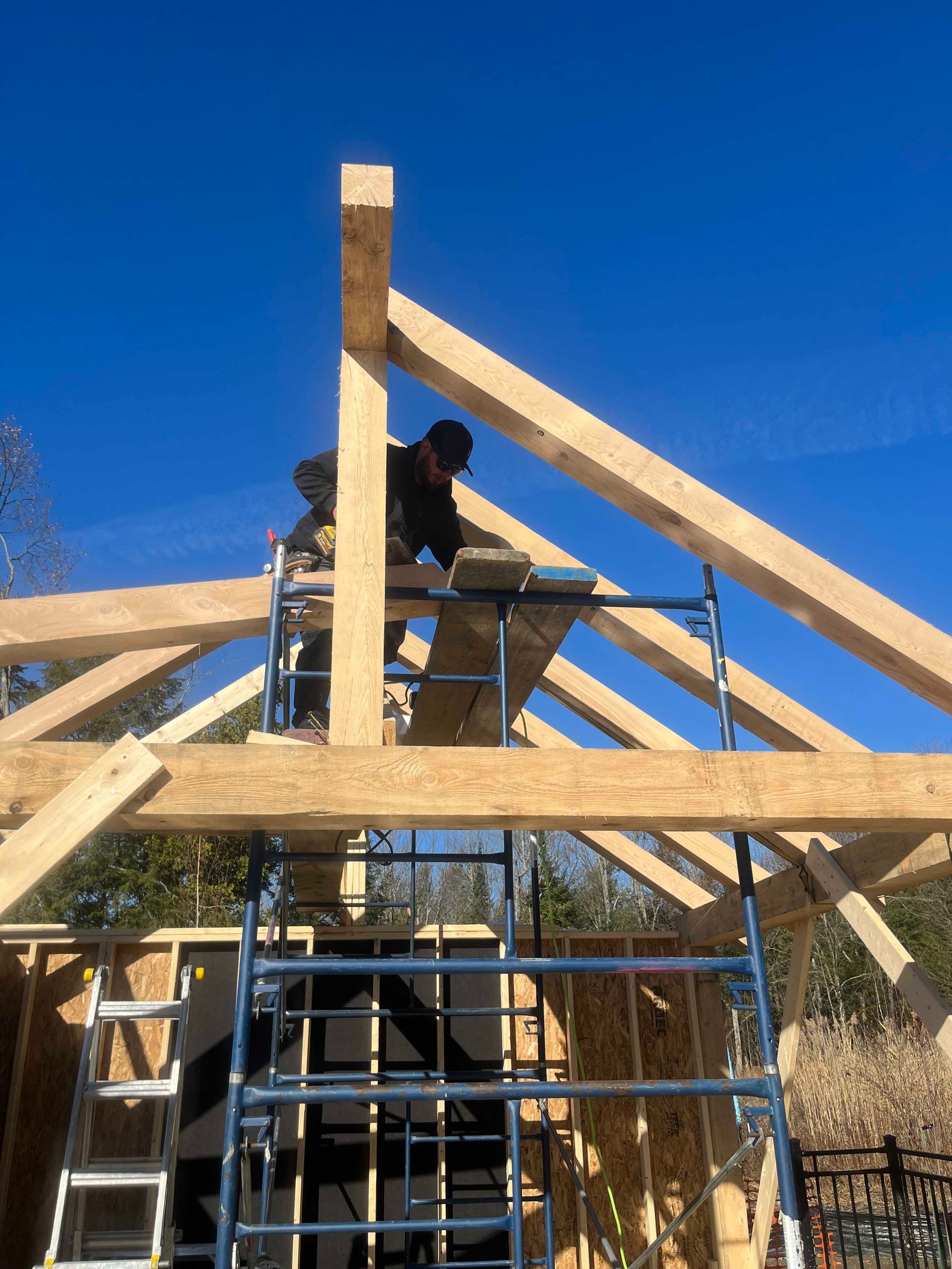 Un ouvrier du bâtiment, perché sur un échafaudage, installe une lourde poutre de toit en bois sur fond de ciel bleu dégagé.