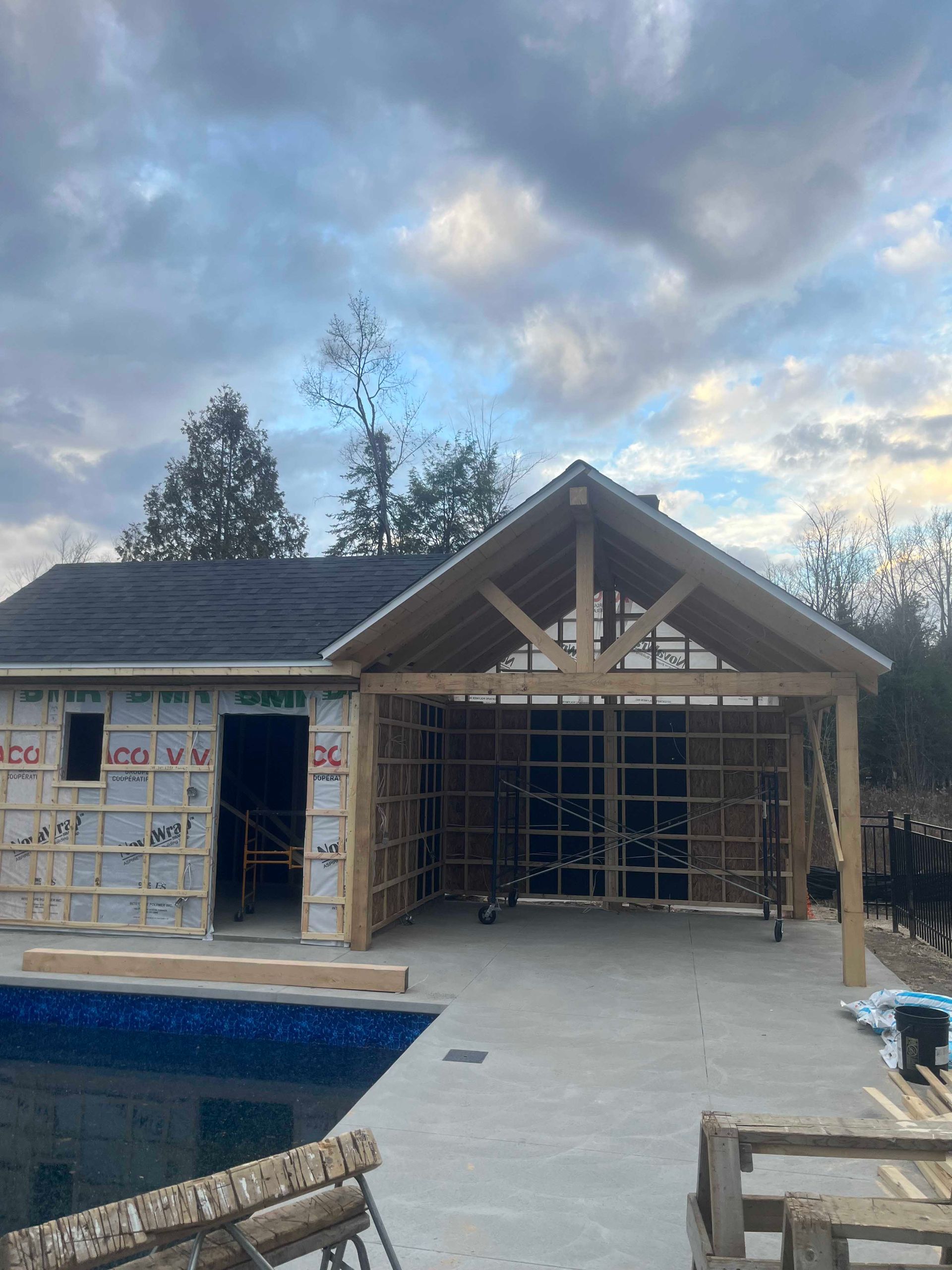 Une structure en bois inachevée, dotée d'un toit en forme de porche, se dresse à côté d'une piscine sous un ciel nuageux.